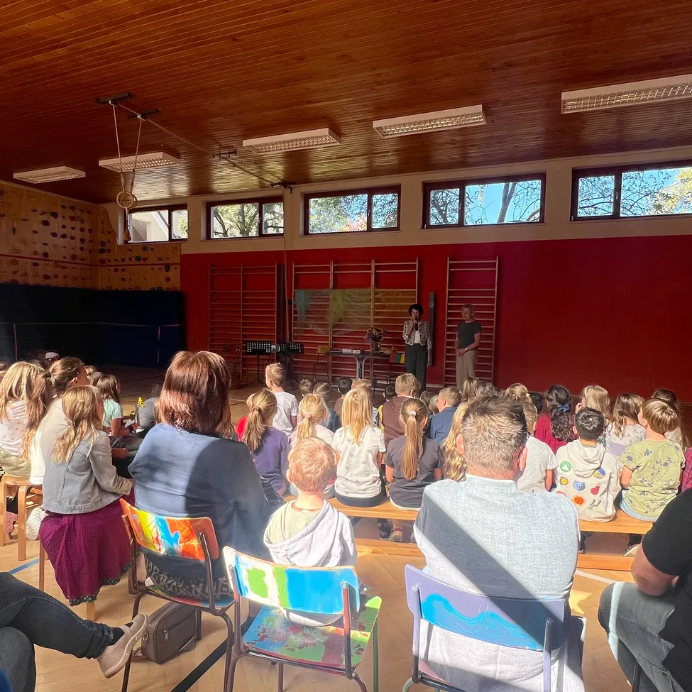 Eine Gruppe von Menschen sitzt in einem Raum mit roten Wänden und einer Holzdecke. Einige sitzen auf Stühlen, andere stehen. Zwei Personen stehen auf der Bühne und eine Frau steht in der Nähe der Tür.
