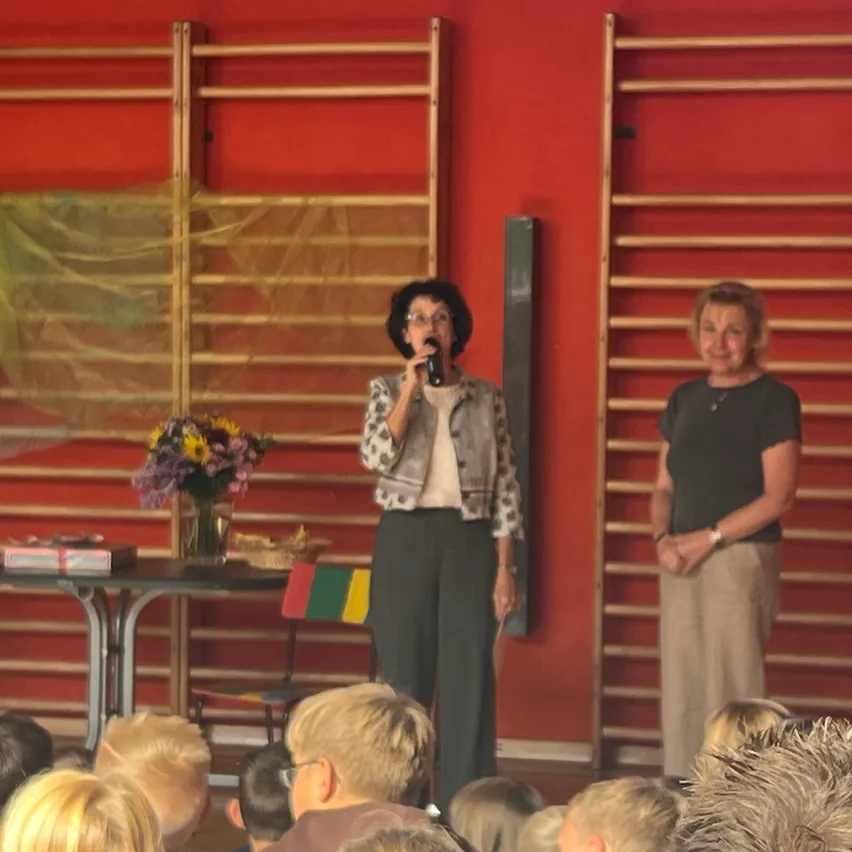 Zwei Frauen stehen auf der Bühne vor einer roten Wand, eine hält ein Mikrofon und spricht, während die andere neben ihr steht. Ein Tisch mit einem Blumenvase und einem Buch steht hinter ihnen. Das Publikum sitzt vor der Bühne.