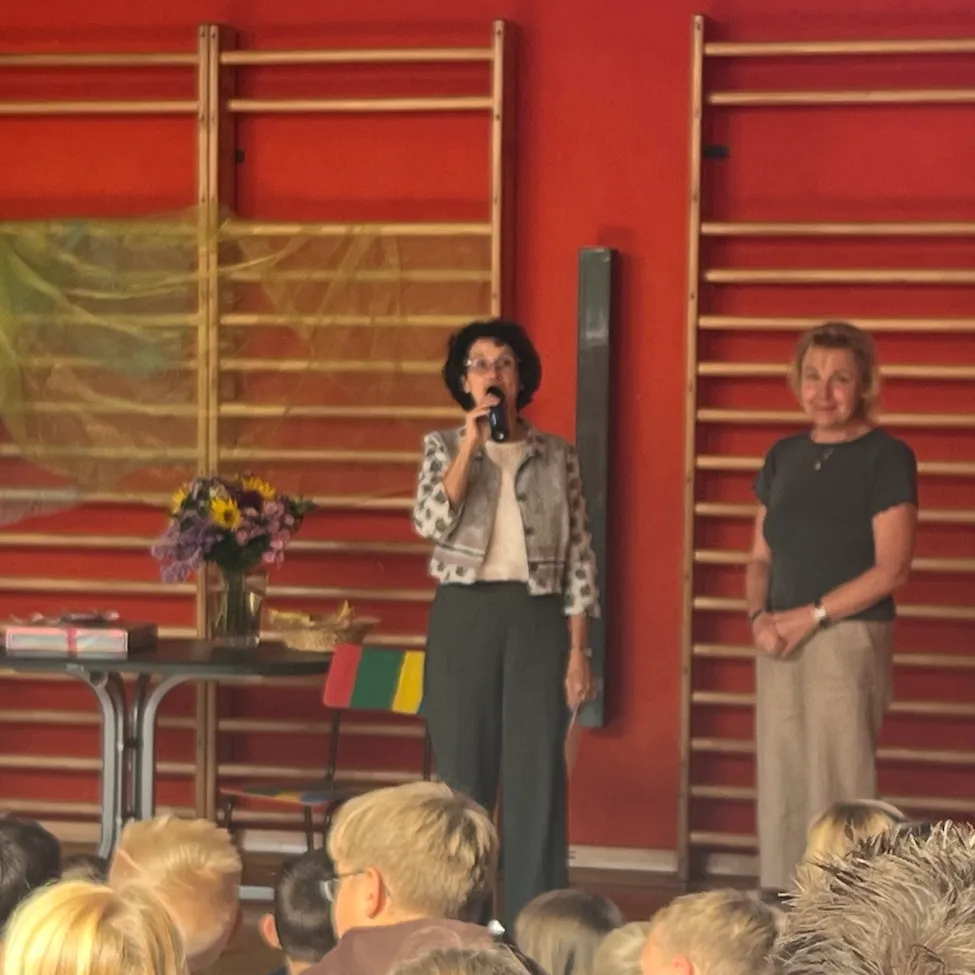 Zwei Frauen stehen auf der Bühne vor einer roten Wand, eine hält ein Mikrofon und spricht, während die andere neben ihr steht. Ein Tisch mit einem Blumenvase und einem Buch steht hinter ihnen. Das Publikum sitzt vor der Bühne.