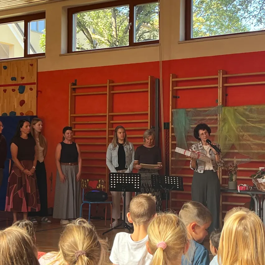 Eine Gruppe von Frauen und Kindern versammelt sich in einem Raum. Die Frauen stehen auf der Bühne, während die Kinder auf dem Boden vor ihnen sitzen. Der Raum hat eine rote Wand und eine Holzwand mit einer Kletterwand.