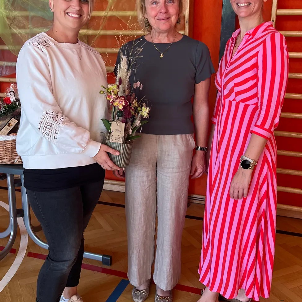 Drei Frauen stehen zusammen in einem Raum mit Holzböden und einer orangefarbenen Wand. Die Frau auf der linken Seite hält einen Blumenstrauß, die Frau in der Mitte hält eine kleine Schachtel und die Frau auf der rechten Seite trägt ein rosa Kleid.