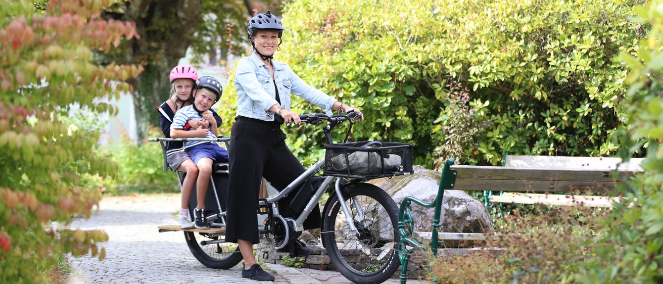 Eine Frau mit zwei Kindern auf einem Lastenrad in einem Park mit Bäumen und einer Bank.
