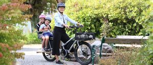 A woman with two children on a cargo bike in a park with trees and a bench.