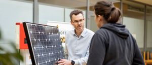 Ein Mann in einem weißen Hemd und mit Brille steht neben einem Solarpanel und unterhält sich mit einer Frau in einer schwarzen Kapuzenjacke.