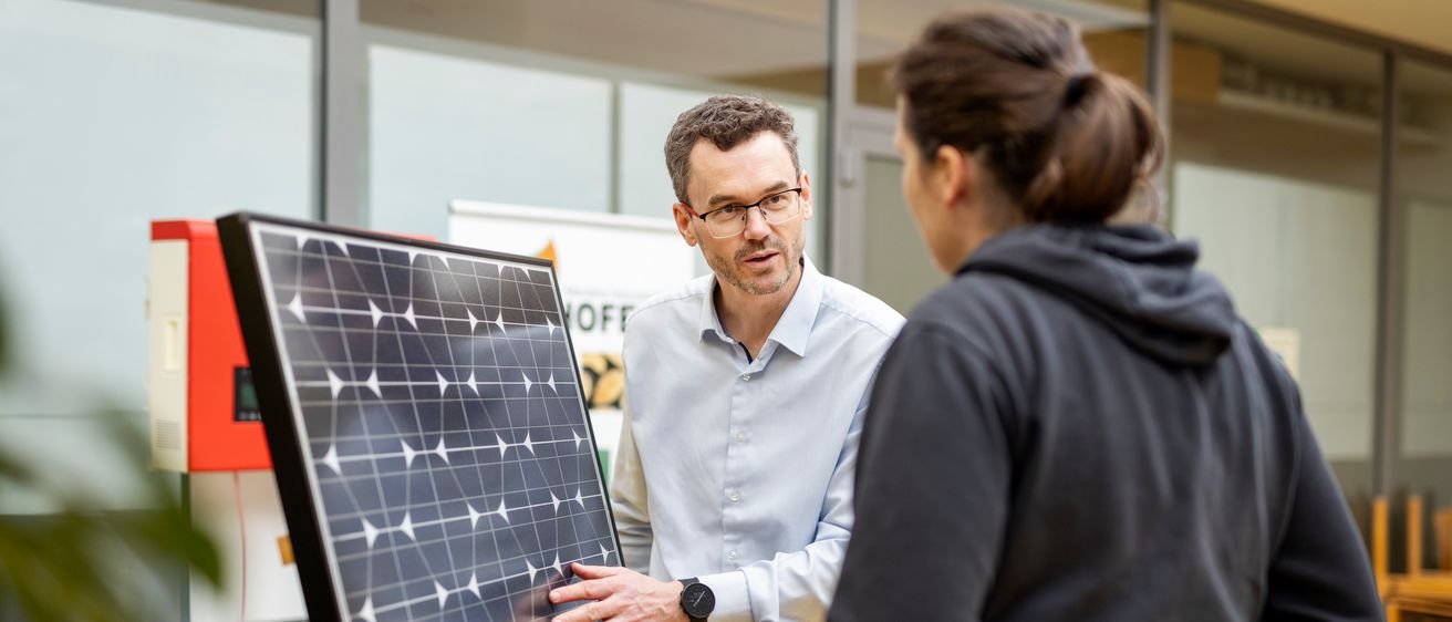 Ein Mann in einem weißen Hemd und mit Brille steht neben einem Solarpanel und unterhält sich mit einer Frau in einer schwarzen Kapuzenjacke.