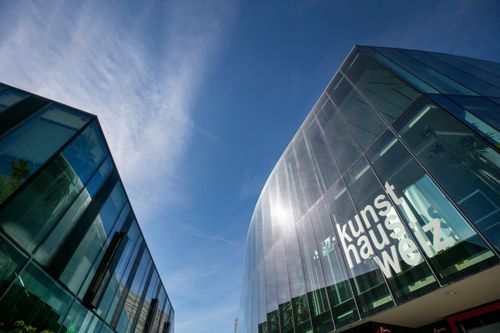 Ein modernes Glasgebäude mit dem Wort 'Kunsthaus' darauf, unter einem klaren blauen Himmel.