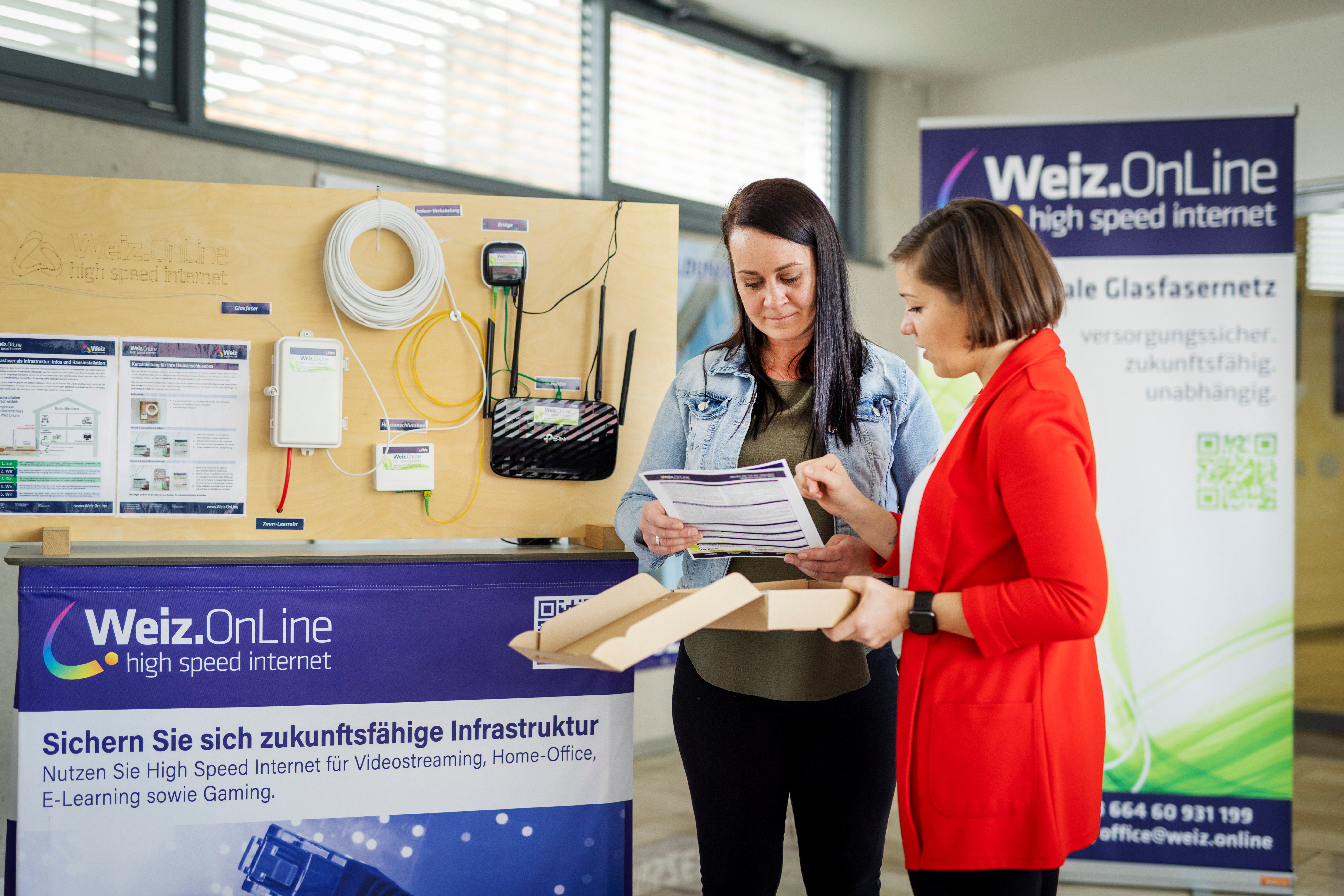 Zwei Frauen stehen vor einem Ausstellungsbrett mit verschiedenen Kabeln und Geräten. Eine Frau hält ein Papier und schaut darauf, während die andere Frau das Papier ansieht. Das Ausstellungsbrett hat ein Schild mit der Aufschrift 'Online'.