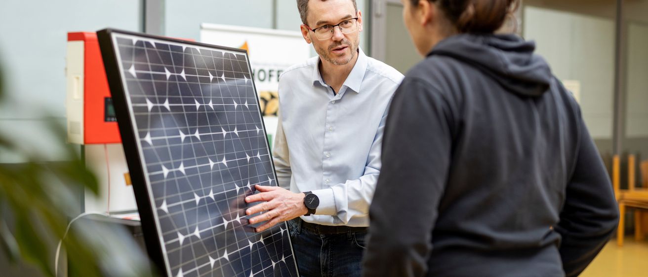 Ein Mann mit Brille und einer Uhr erklärt einer Frau in einer Kapuzenjacke etwas über ein Solarpanel.