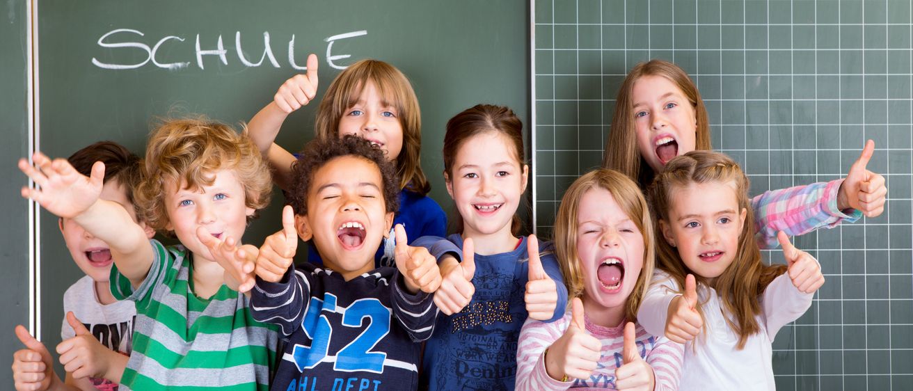 Eine Gruppe von Kindern mit erhobenen Daumen vor einer Tafel, auf der 'SCHULE' steht.