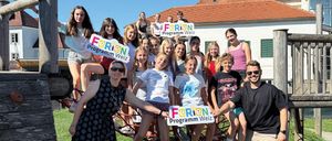 Eine Gruppe von Menschen posiert für ein Foto in einem Outdoor-Spielplatz. Sie halten Schilder, auf denen Ferien Programm Weiz steht.