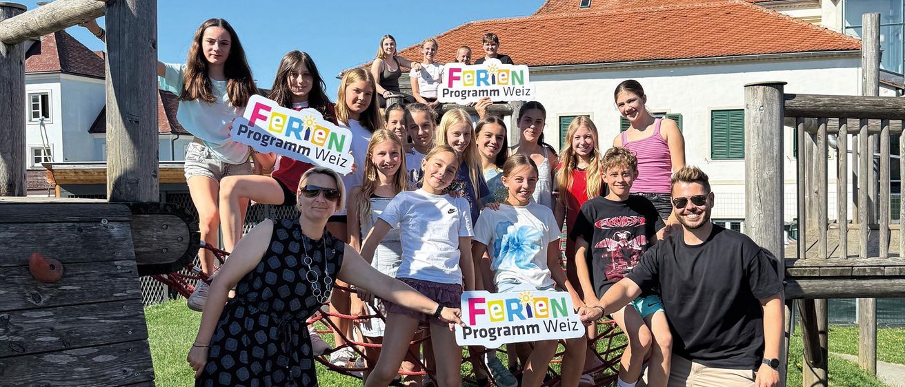 Eine Gruppe von Menschen posiert für ein Foto in einem Outdoor-Spielplatz. Sie halten Schilder, auf denen Ferien Programm Weiz steht.