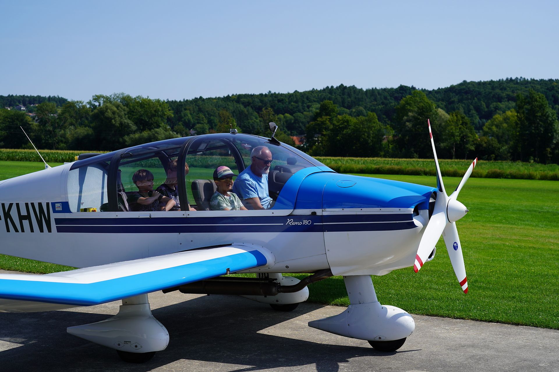 Vier Personen sitzen in einem kleinen blau-weißen Flugzeug mit einem weißen Propeller. Sie sind draußen an einem sonnigen Tag mit Bäumen und Gras im Hintergrund.