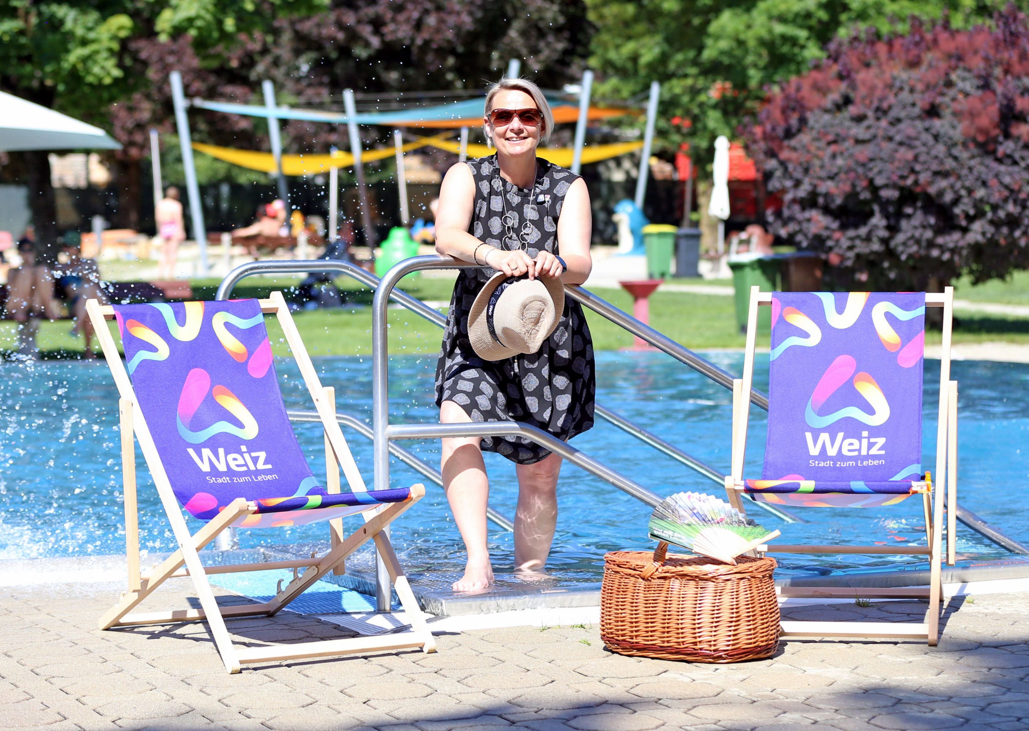Eine Frau in einem Kleid und mit Sonnenbrille steht im Pool, hält einen Hut und lächelt. Zwei farbenfrohe Liegestühle und ein Korb sind in der Nähe.