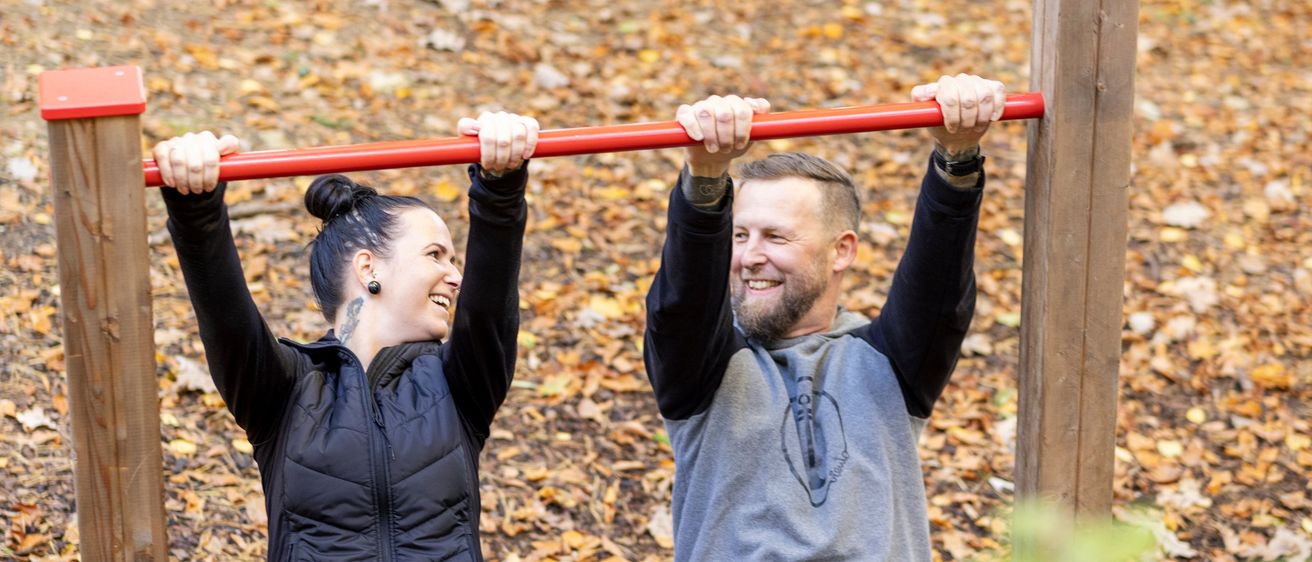 Ein Mann und eine Frau machen Klimmzüge in einem Waldpark. Sie lächeln und scheinen die Übung zu genießen. Der Boden ist mit trockenen Blättern bedeckt.