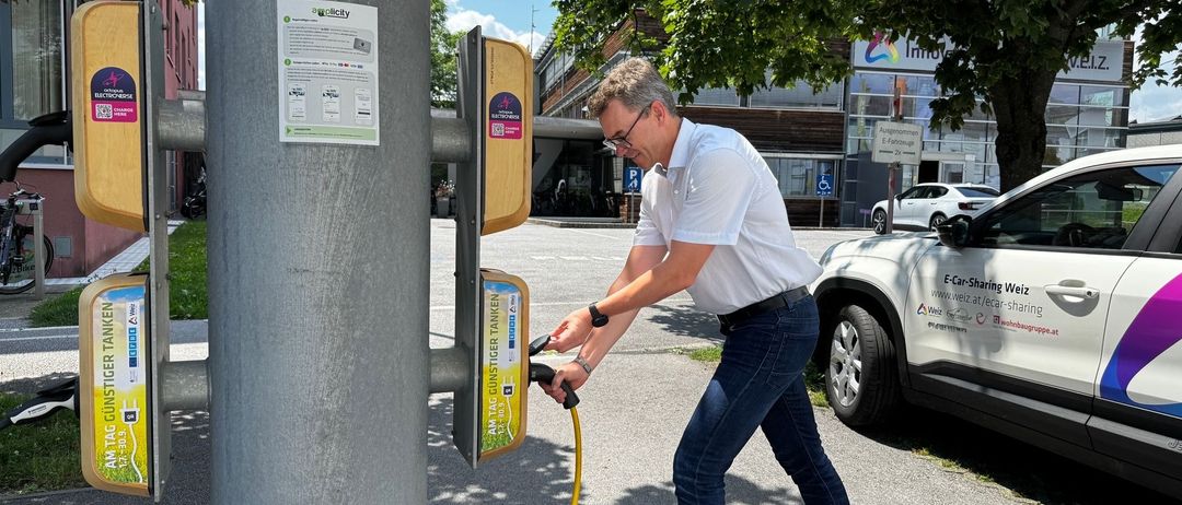 Ein Mann verbindet sein Elektroauto mit einer Ladestation mit einem gelben Kabel. Die Station ist an einem Pfosten mit einem gelben Schild montiert. Dahinter ist ein Gebäude zu sehen.