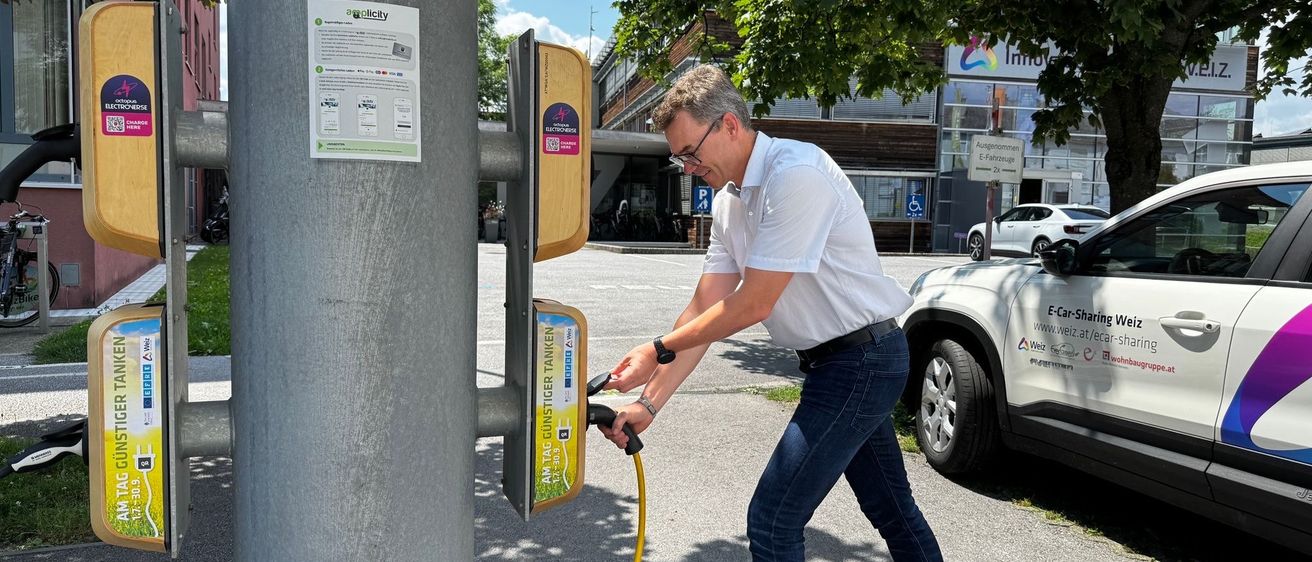 Ein Mann verbindet sein Elektroauto mit einer Ladestation mit einem gelben Kabel. Die Station ist an einem Pfosten mit einem gelben Schild montiert. Dahinter ist ein Gebäude zu sehen.