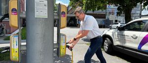 Ein Mann verbindet sein Elektroauto mit einer Ladestation mit einem gelben Kabel. Die Station ist an einem Pfosten mit einem gelben Schild montiert. Dahinter ist ein Gebäude zu sehen.