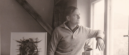 A man stands in a room, looking out the window. He wears a sweater with paint stains. Beside him is a canvas with abstract art. A potted plant and paintbrushes are on the windowsill.