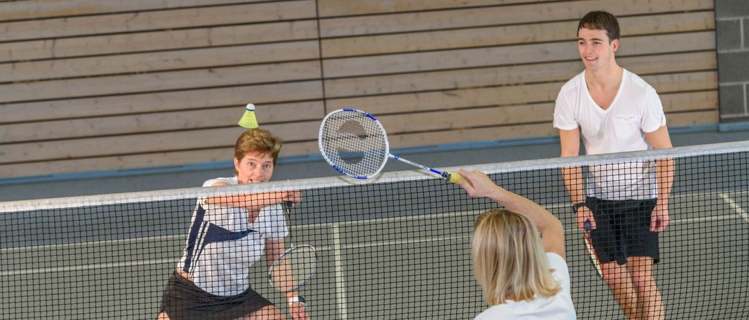 Drei Menschen spielen Badminton in Innenräumen. Ein Mann und eine Frau schlagen einen Shuttlecock, während ein anderer Mann zuschaut.