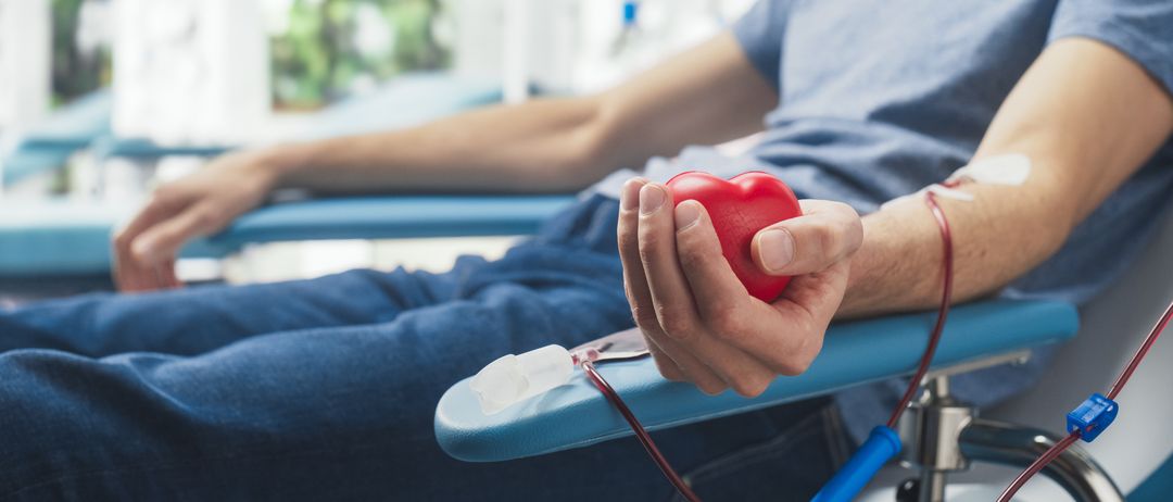 Ein Mann spendet Blut in einem Krankenhaus. Er hält eine rote Herz in der Hand, mit einem Blutschlauch angeschlossen.