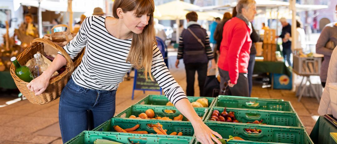 Eine Frau betrachtet Gemüse auf einem Bauernmarkt. Sie hält einen Korb und schaut sich Karotten und Blattgemüse vor sich an. Andere Käufer sind im Hintergrund zu sehen.