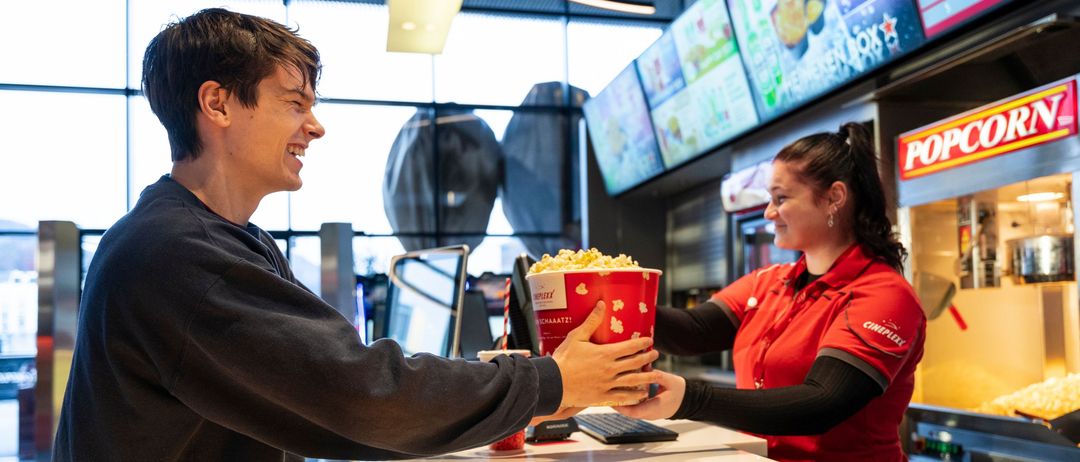 Ein Mann lächelt, während er einer Frau in einer roten Uniform eine Popcorntüte überreicht, an einem Essensschalter.