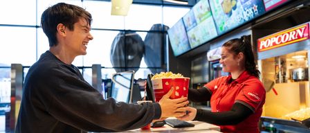 Ein Mann lächelt, während er einer Frau in einer roten Uniform eine Popcorntüte überreicht, an einem Essensschalter.