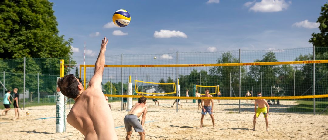Ein Mann springt, um den Volleyball zu spiken, während andere zuschauen und auf einem sandigen Beachvolleyballplatz spielen.
