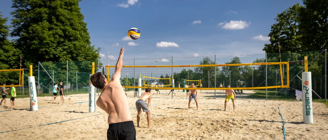 Eine Gruppe von Menschen spielt Volleyball auf einem sandigen Spielfeld. Ein Spieler ist kurz davor, den Ball zu schlagen. Das Spielfeld ist von einem Netz umgeben, und im Hintergrund befinden sich Bäume und Gebäude.