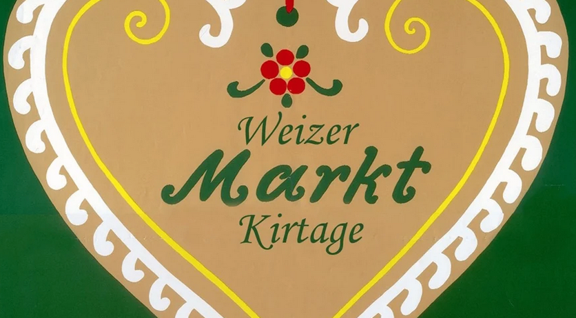 Ein herzförmiges Schild mit 'Weizer Markt Kirtage' in Grün und Gelb, das von einem roten Band an einem grünen Hintergrund mit festlichen Elementen hängt.