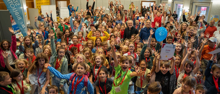 Eine große Gruppe von Kindern, einige mit Abzeichen, jubelt und hebt die Hände. Ein Mann in einem blauen Mantel steht in der Mitte. Ein Banner hängt an der Wand im Hintergrund.