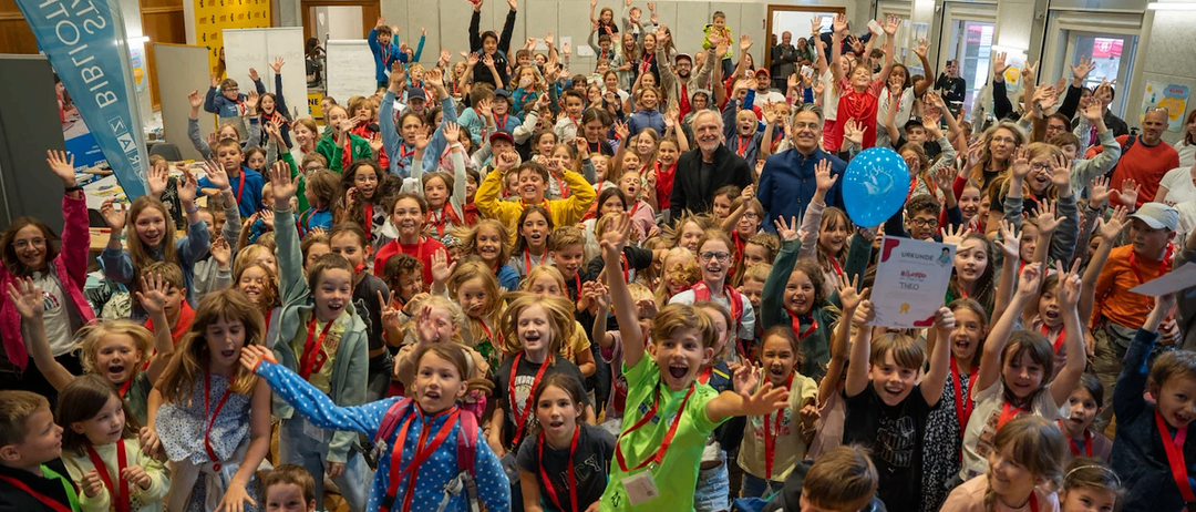 Eine große Gruppe von Kindern, einige mit Abzeichen, jubelt und hebt die Hände. Ein Mann in einem blauen Mantel steht in der Mitte. Ein Banner hängt an der Wand im Hintergrund.