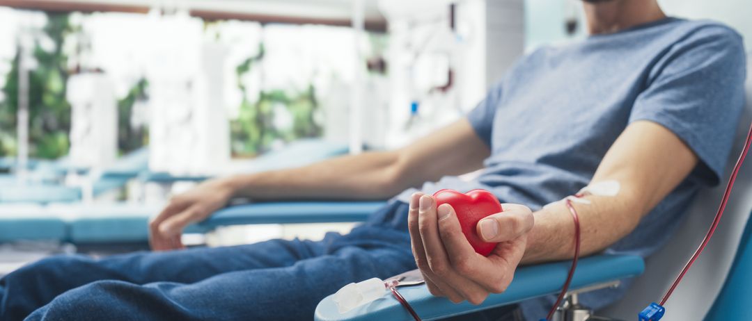 Ein Mann im Krankenhausbett hält ein rotes Herz und ist an eine Blutspende-Kanüle angeschlossen.