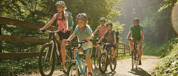 Eine Gruppe von Kindern und Erwachsenen fährt auf einem Waldweg mit Fahrrädern, trägt Helme und Rucksäcke.