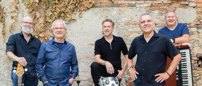 Fünf Männer stehen vor einer Ziegelwand, lächeln und posieren für ein Foto. Sie halten Musikinstrumente in den Händen, darunter eine Gitarre, Trommeln und ein Keyboard.
