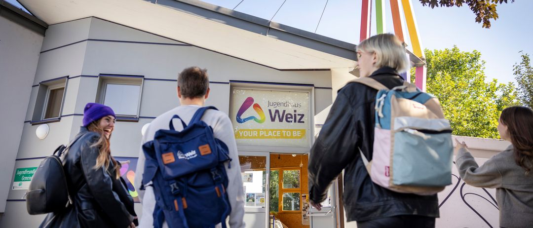 Drei Personen mit Rucksäcken gehen vor einem Gebäude mit dem Schriftzug 'Jugendhaus Welz'. Das Gebäude hat einen bunten Bogen über dem Eingang.