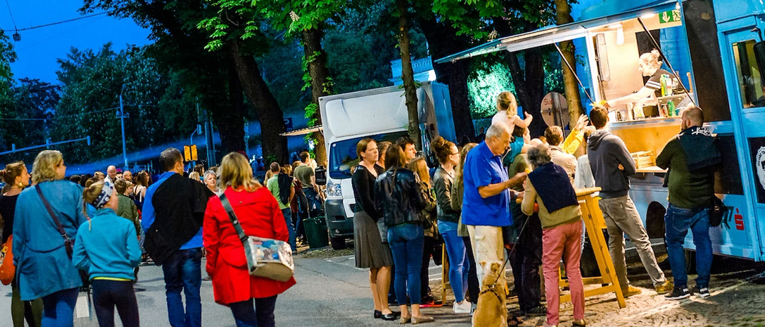 Eine Gruppe von Menschen versammelt sich um einen Food-Truck auf einer Straße bei Nacht. Der Food-Truck ist blau, und eine Frau steht darin. Die Menschen stehen vor dem Truck, und einige halten Taschen. Die Straße ist von Bäumen und Straßenlaternen umgeben.