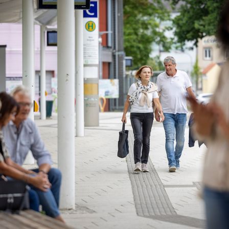 Eine urbane Straßenszene mit einem Paar, das an sitzenden Personen vorbei an einer Bushaltestelle geht. Bäume und Gebäude sind im Hintergrund.