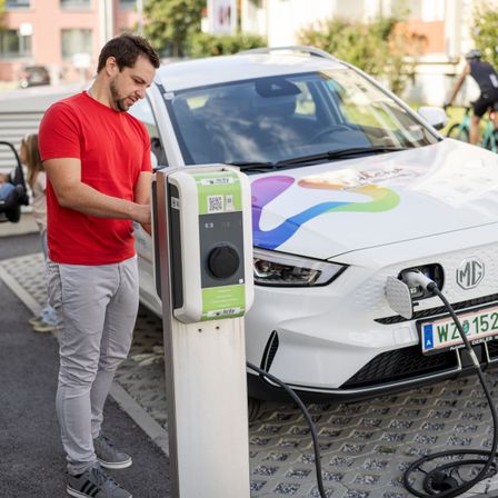 Ein Mann steht an einer Ladestation für Elektrofahrzeuge und schließt sein Auto an. Er trägt ein rotes T-Shirt und graue Hose. Im Hintergrund ist ein Fahrradfahrer zu sehen.