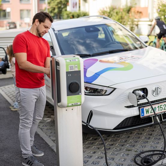 A man stands at an electric vehicle charging station, plugging in his car. He wears a red shirt and gray pants. A bicycle rider is in the background.