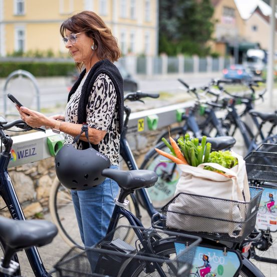 An elderly woman stands at a bike rack, holding her phone and looking at it, wearing a leopard print top, jeans, and a black helmet. Several bicycles are parked, including one with a bag of vegetables on the front.