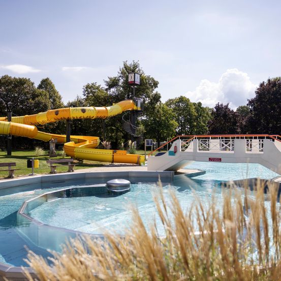 A water park with a yellow slide and a pool, surrounded by green trees and a bridge.