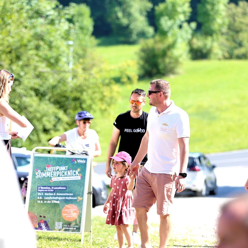 Eine Familie geht auf einem Rasengelände in der Nähe einer Straße. Der Vater hält die Hand der Tochter. Dahinter steht ein Schild mit einem grünen Plakat. Im Hintergrund sind mehrere geparkte Autos.