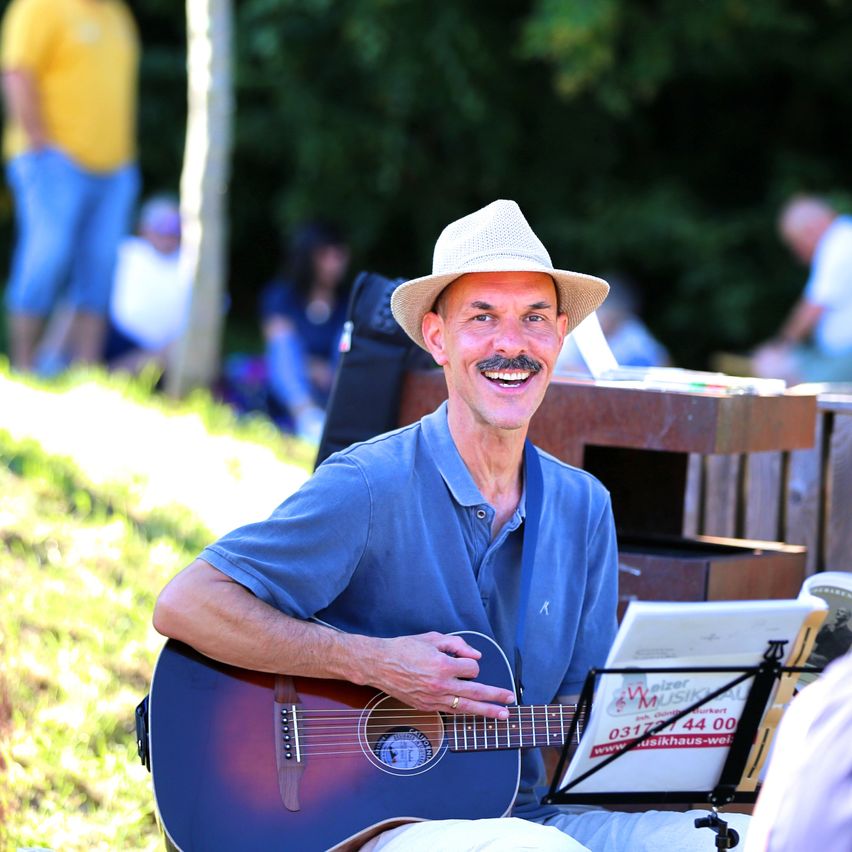 Ein Mann mit Hut und Schnurrbart lächelt, während er die Gitarre spielt. Er sitzt auf dem Rasen.