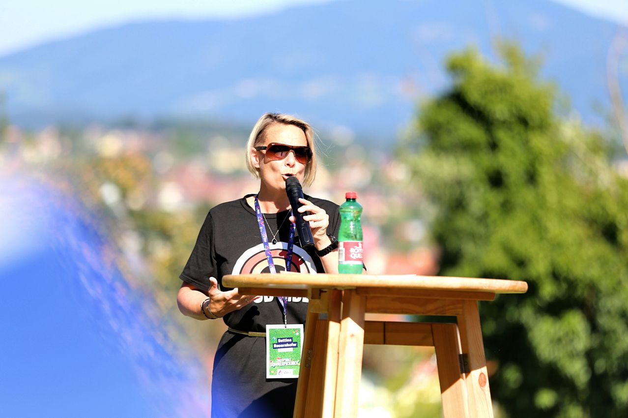 Eine Frau mit Sonnenbrille und schwarzem T-Shirt steht an einem Holzpodium und hält ein Mikrofon. Eine grüne Flasche steht auf dem Podium und eine verschwommene Ansicht einer Stadt und Bäume ist hinter ihr.