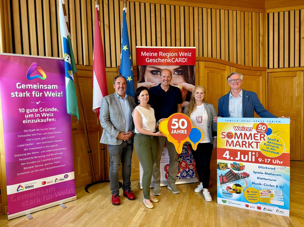 Fünf Menschen stehen lächelnd vor einer Holzwand. Drei Banner stehen dahinter. Zwei Personen halten ein großes Schild, auf dem 50 Jahre Sommer Markt steht.