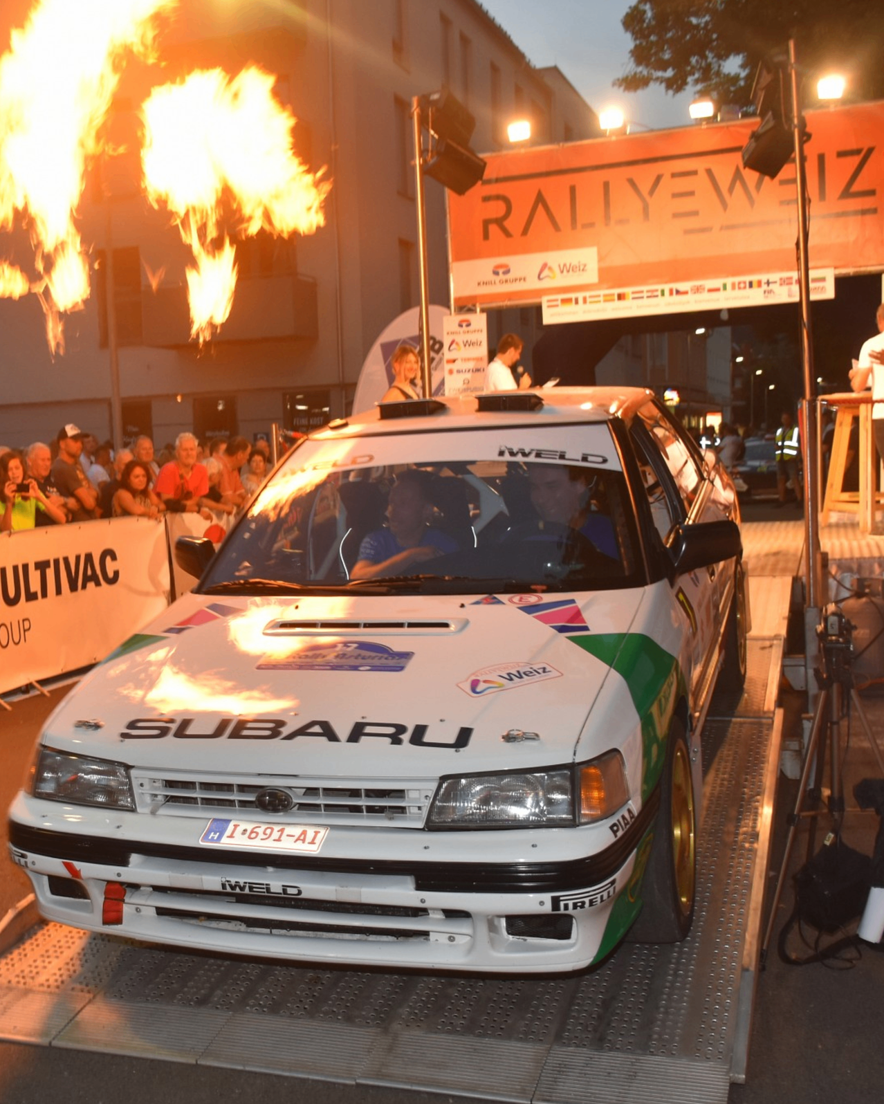 Ein Subaru Rallye-Auto wird bei einer Veranstaltung mit einem Publikum ausgestellt. Das Auto hat das Kennzeichen I-691-AI. Eine Person steht in der Nähe eines Podiums, und es gibt Lautsprecher und Lichter auf der Bühne.