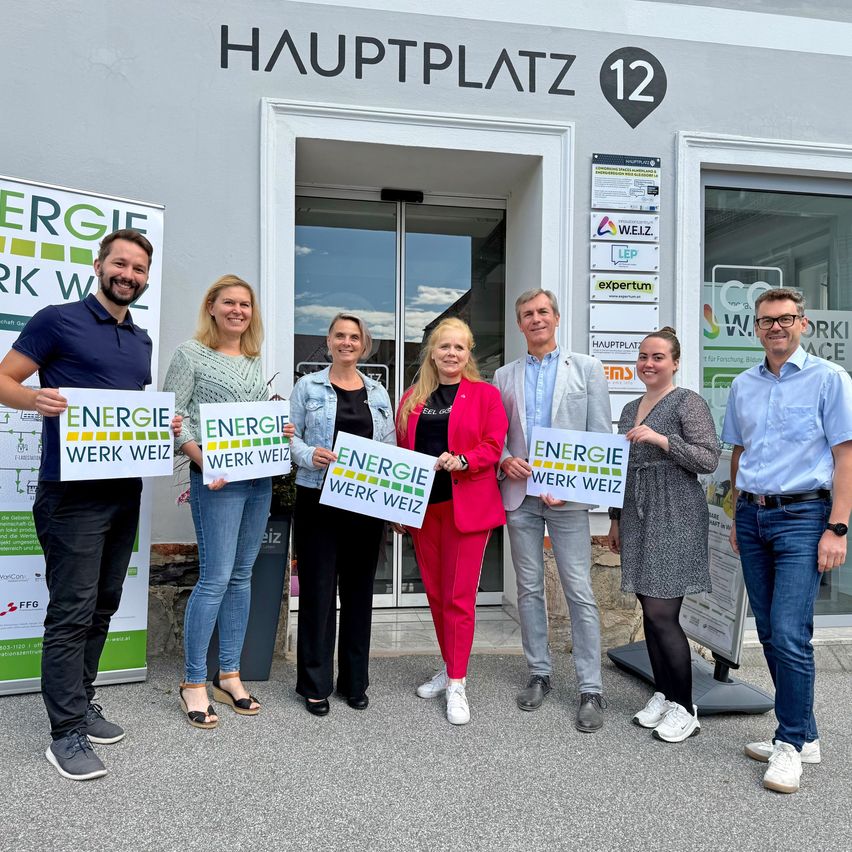 Eine Gruppe von Menschen steht vor einem Gebäude mit der Aufschrift 'Hauptplatz 12'. Sie halten Schilder mit der Aufschrift 'Energie Werk Weiz'.