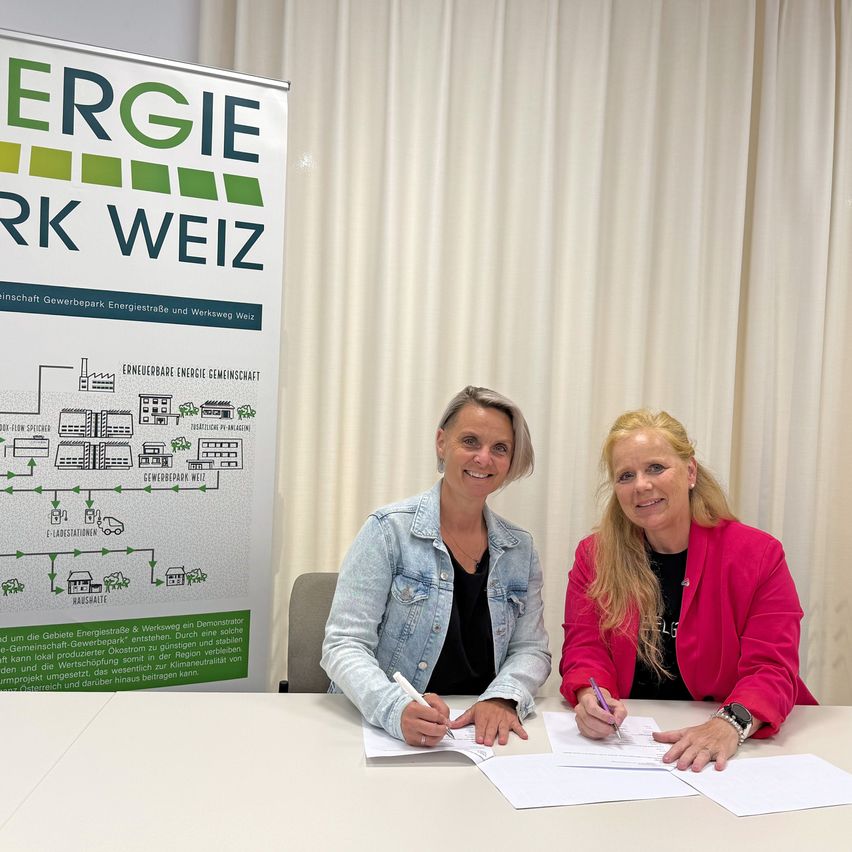 Zwei Frauen sitzen an einem Tisch und unterschreiben Dokumente. Eine trägt eine Jeansjacke, die andere einen rosa Blazer. Hinter ihnen steht ein Banner mit der Aufschrift 'Energiepark Weiz'. Papiere und Stifte liegen auf dem Tisch, und ein Hintergrund aus weißen Vorhängen ist sichtbar.