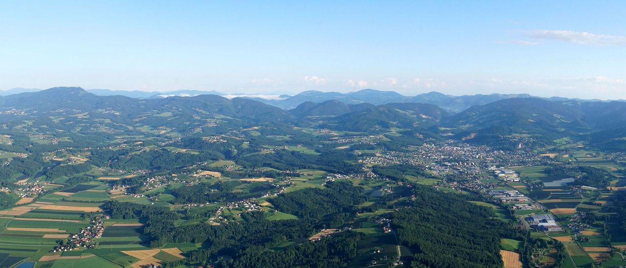 Luftaufnahme eines Tals mit üppigem grünen Bäumen und Häusern, umgeben von Bergen unter einem klaren blauen Himmel.
